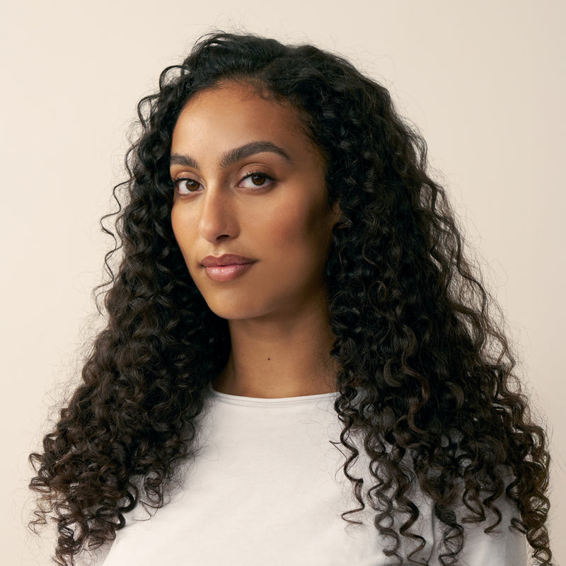 Woman with long, defined shiny curly black hair wearing a white shirt against a beige background