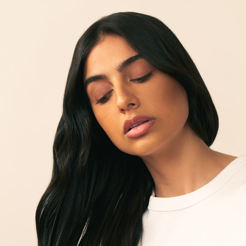 Woman with long shiny, healthy looking black hair wearing a white top against a beige background