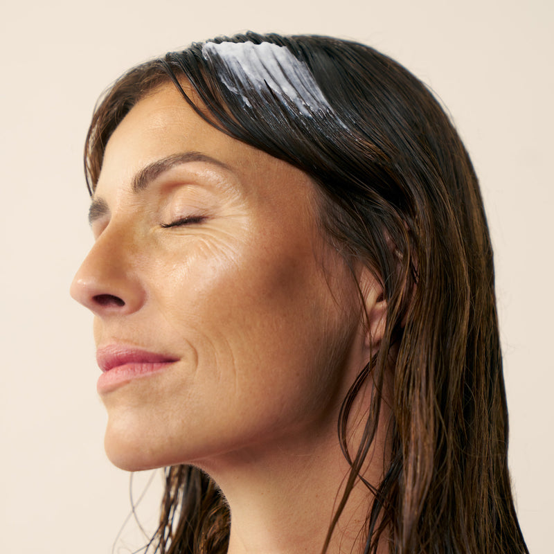Woman with wet hair and a white hair mask on her head against a beige background