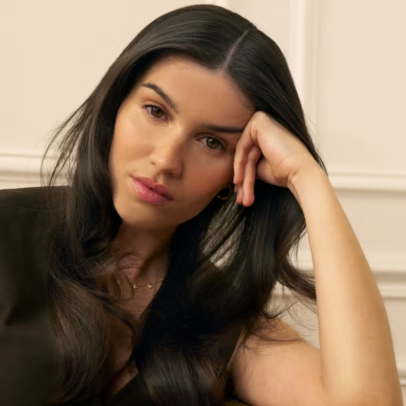 Woman with long dark shiny hair resting her head on her hand against a neutral background 
