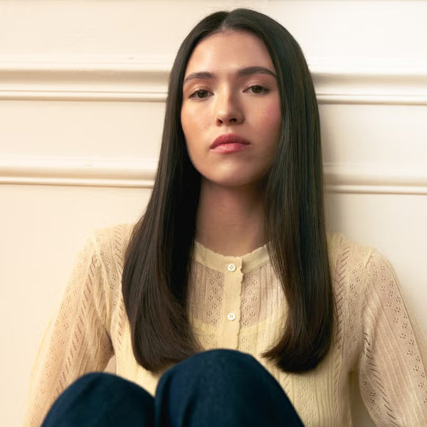 Woman with glossy hydrated dark hair sitting against a light-colored wall wearing a beige sweater and white blouse.