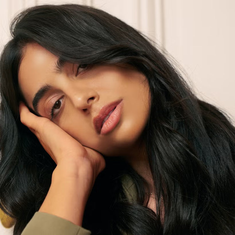 Woman with long glossy black hair resting her head on her hand against a neutral background